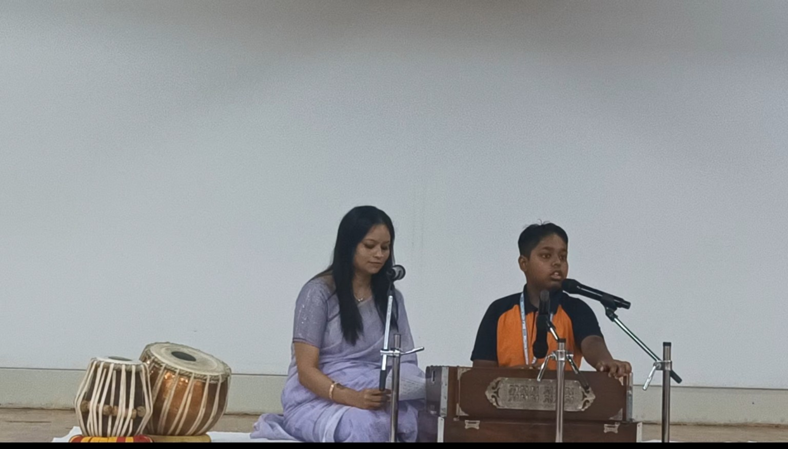 Mamta Gangwal beside Shivoham as he sings at a microphone with a harmonium